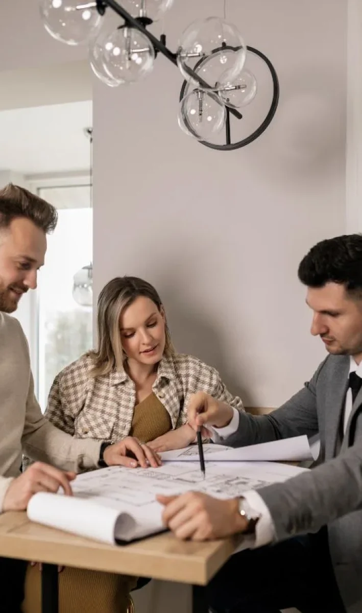 Three people are gathered around a high table in a bright room, examining architectural blueprints. One person, wearing a suit, points at the plans with a pen, while the other two, casually dressed, attentively look on.