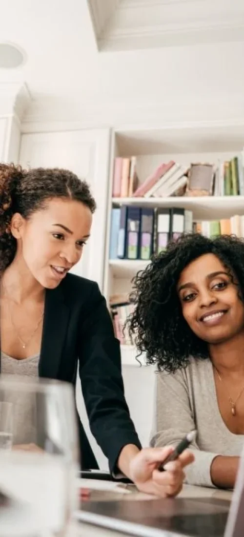 Two women are working together in a modern office. One woman, with curly hair and wearing a blazer, is standing and pointing at a laptop screen. The other woman, with curly hair as well and wearing a sweater, is sitting and smiling. Bookshelves are in the background.