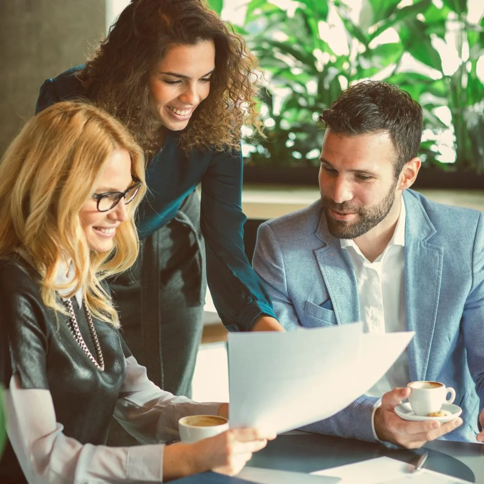 Three colleagues at a cafe table, with two women and one man. One woman is standing and engaging with the seated woman and man who are looking at a document. Both seated individuals have coffee cups. The background features green plants.