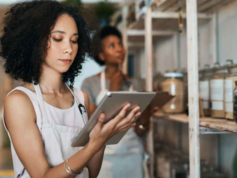 A woman with curly hair, wearing a light-colored apron, uses a tablet in a store. Behind her, another person examines jars of goods on shelves. The store has a bright, organized, and neatly arranged setting.