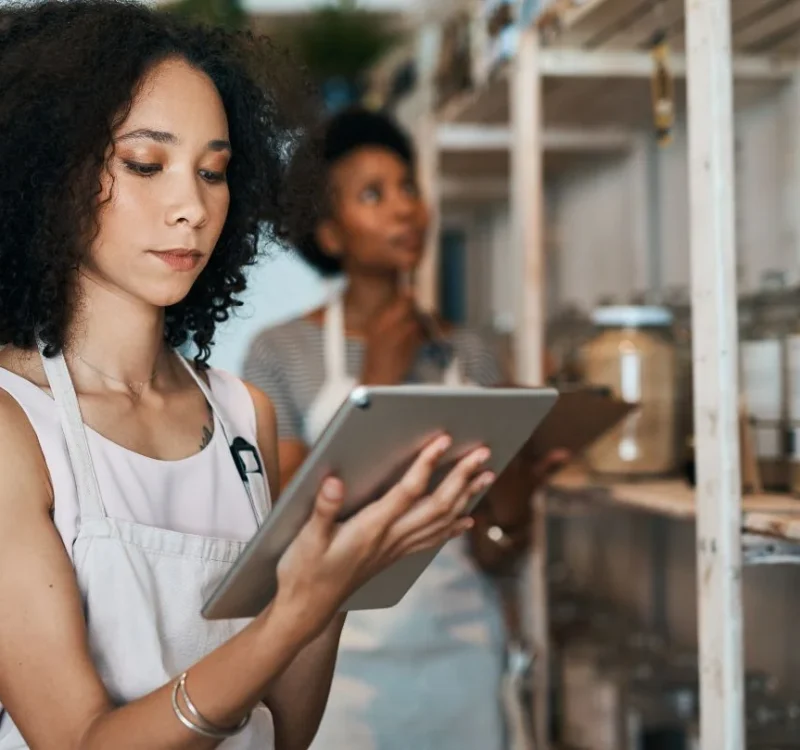 A woman with curly hair, wearing a light-colored apron, uses a tablet in a store. Behind her, another person examines jars of goods on shelves. The store has a bright, organized, and neatly arranged setting.