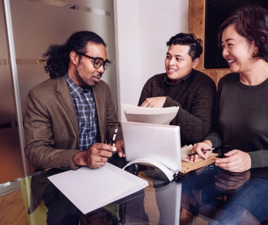 Three colleagues sit around a glass table and discuss documents. One holds a paper, another points at it, and the third person laughs while writing. A laptop and notepad are on the table. They are in an office setting with modern furnishings.