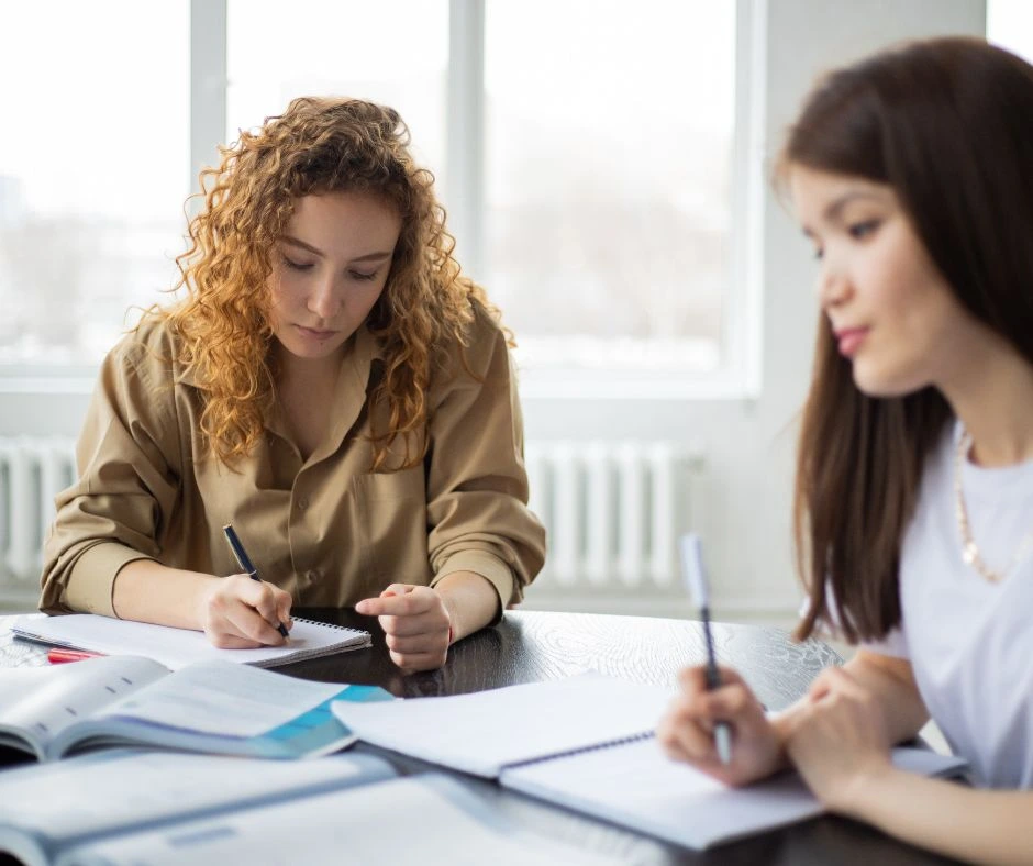 Two women are seated at a table, focused on writing in notebooks, with various open books and papers spread out before them. The woman on the left has curly hair and is wearing a beige shirt, while the woman on the right has long straight hair and is wearing a white shirt.