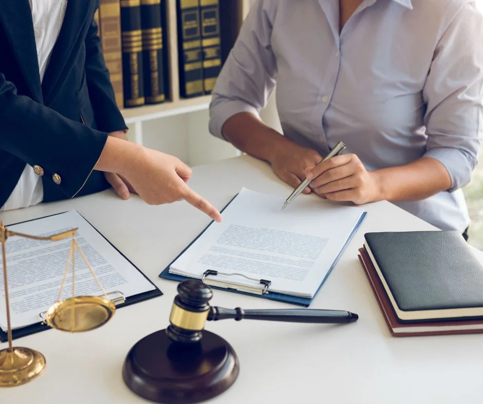 Two people are sitting at a desk, reviewing documents. One person is pointing at a document, while the other holds a pen. On the desk are a gavel, a pair of scales, a stack of books, and a clipboard. Shelves with books are in the background.