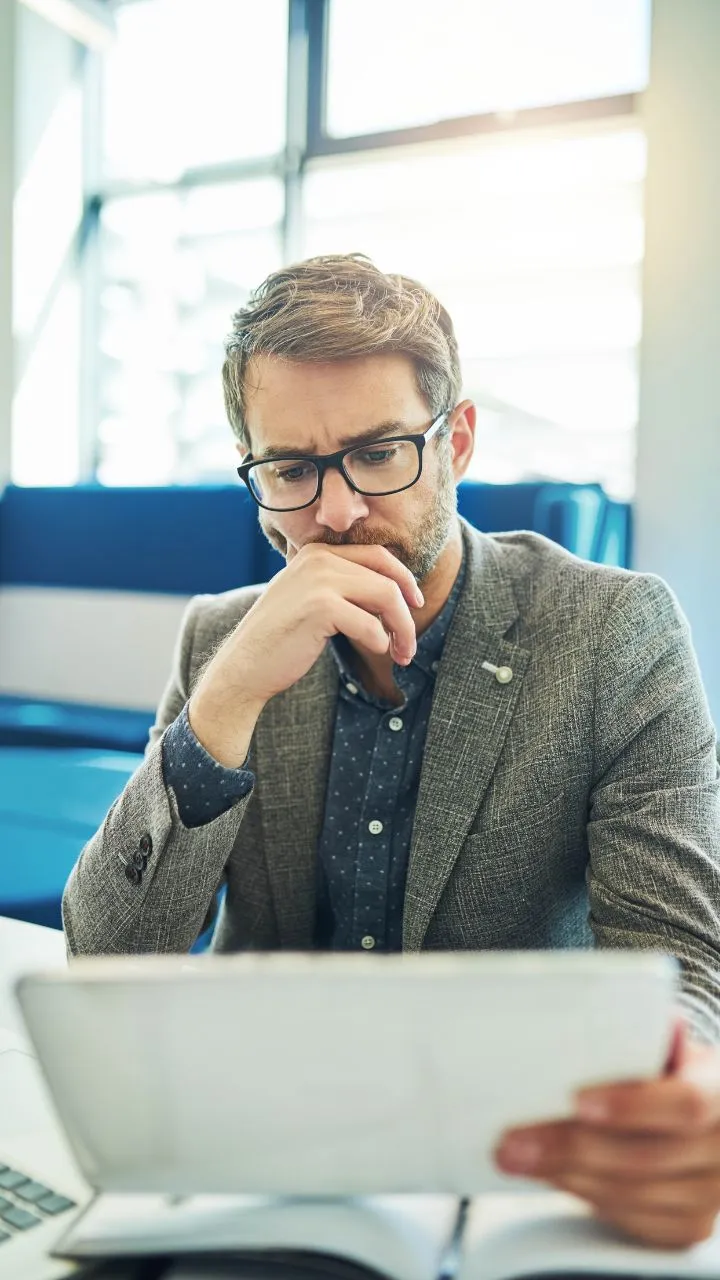 A man with glasses and a beard sits indoors, reading a document with a thoughtful expression. He is dressed in a grey blazer and a blue shirt with white polka dots. The background shows a bright office space with large windows and blue furniture.