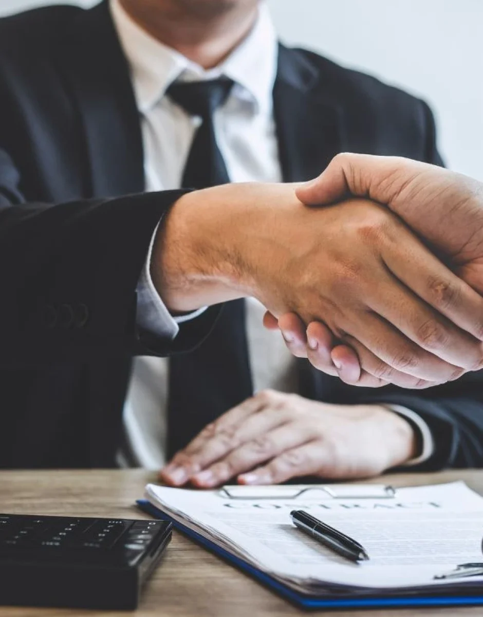 Two people are shaking hands over a desk, with documents, a calculator, and a pen organized neatly on top. One person is wearing a suit and tie, while the other person's hand is visible. The scene appears professional, suggesting a business agreement.