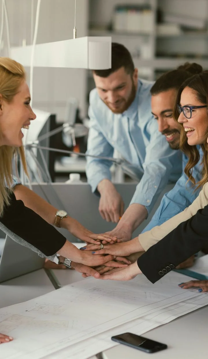 A group of four colleagues stand around a table in an office, placing their hands together in the center in a gesture of teamwork. They are smiling and appear enthusiastic. The table has some documents and a smartphone on it.