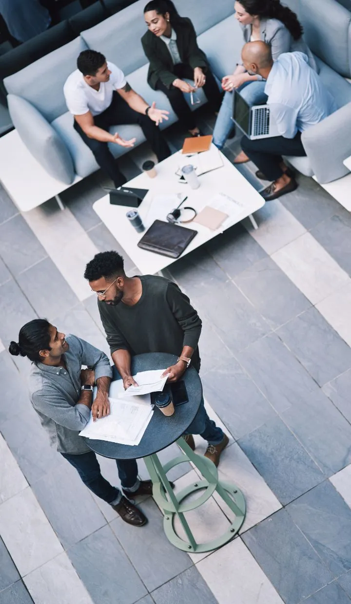 Top view of a modern office space showing two groups of people collaborating. One group is sitting on sofas having a discussion with laptops and notebooks, while a pair stands around a high table, engaged in conversation over documents.