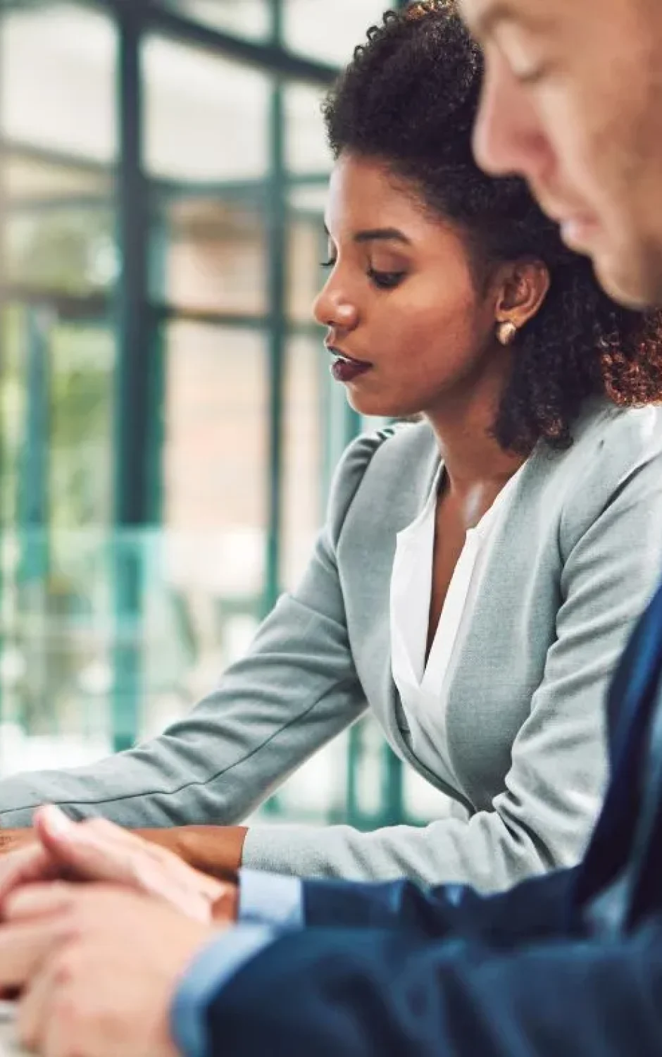 A woman and a man are sitting at a conference table in a modern office. The woman, wearing a light gray blazer, is writing in a notebook, while the man, in a dark suit, looks at a tablet. Both appear focused on their tasks.